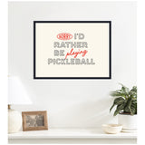 Framed wall art with the text 'SORRY I'D RATHER BE playing PICKLEBALL' in black and red fonts on a cream background, hanging above a white surface with lamp and plants.