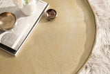 Close-up of a round coffee table with a textured golden surface and thin raised edge, topped with stacked hardcover books and small metallic bowls, on a soft white rug.