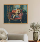 Framed painting of a lion lounging on a red tufted armchair with a deep teal and gold abstract background, hanging on a beige wall above a gray sofa and vase.