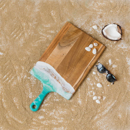 Wooden resin charcuterie board with a turquoise handle on sand, accompanied by sunglasses and a coconut.