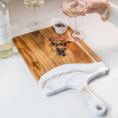 Person holding a glass of white wine with a resin charcuterie board and bottle on a countertop.