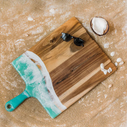 Wooden charcuterie serving tray with a turquoise wave design on sand, accompanied by sunglasses and a coconut.