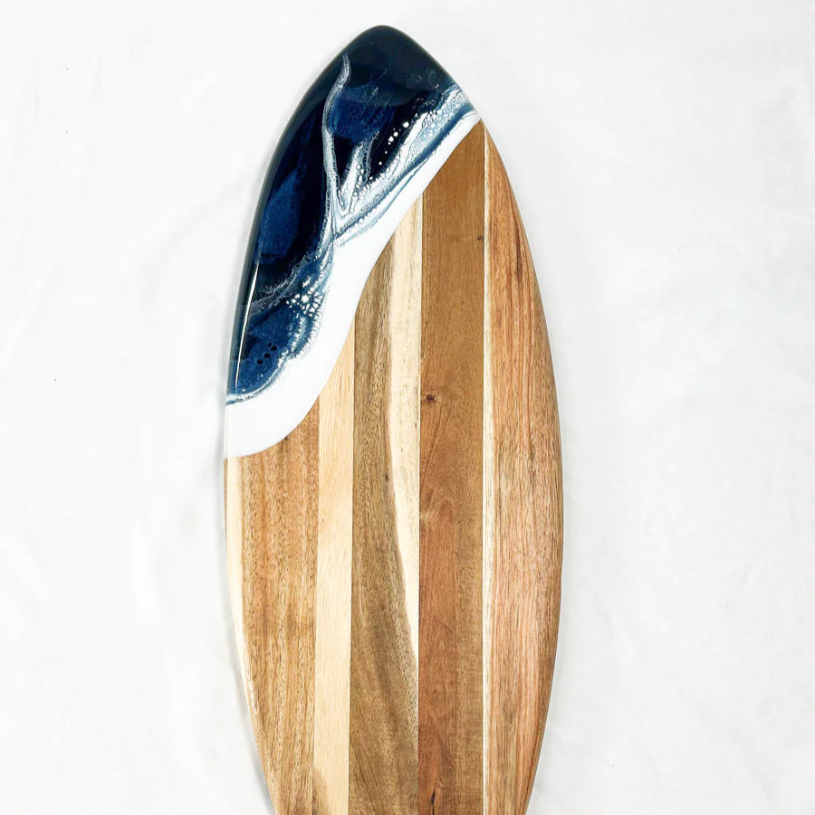 Wooden surfboard-shaped charcuterie board with blue resin accent, holding cheese and fruit on a patio table
