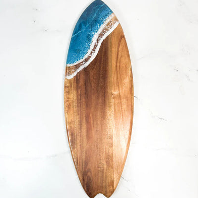 Wooden surfboard-shaped charcuterie board with blue resin accent, holding cheese and fruit on a patio table