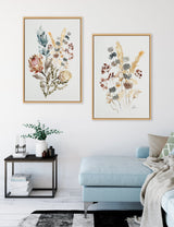 Two framed watercolor paintings of wildflowers in muted tones of green, yellow, and brown hanging above a light blue sectional sofa in a bright living room.