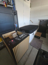 Rectangular cold plunge tub with black insulated cover next to a vertical-striped chiller unit labeled LUX in a plain utility room with concrete floor and white walls.