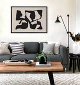 Living room with dark gray sofa adorned with gray and striped pillows, a wood coffee table with black decor and a plant, and a black framed abstract canvas with distorted black shapes on tan background.
