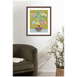 Framed artwork of colorful snowdrop flowers with pink, white, and blue petals in a striped pot on a mustard green background hung above an olive green armchair and woven basket plant.