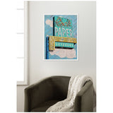 Framed canvas art print of a vintage-style sign reading 'Rock Paper Scissors' in bold teal, yellow, and beige tones with cloud-like shapes on a blue background, hanging above a dark green armchair draped with a white knitted throw