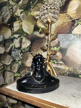 Black bust statue of Adiyogi with detailed facial features and hair, placed on a circular base beside a gold-toned leaf and crystal decorative candle holder against stone-patterned wallpaper.
