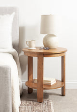 Round wooden side table with lower shelf holding two books, topped with beige ceramic table lamp, glasses, and a white mug beside a light gray upholstered bed.