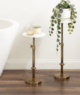 Two adjustable brass stands with round white marble tops on dark wood floor near white wall, one holding a trailing potted plant, the other with glass and candle.