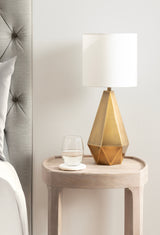 Geometric faceted gold lamp with a cylindrical white shade on a light wood round side table next to a bed with tufted gray headboard and white linens.