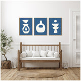 Three framed abstract ceramic shapes in white on blue backgrounds, arranged horizontally above a wooden bench with white cushions in a light, minimalist room.