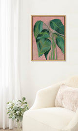 Framed canvas painting of large green Alocasia leaves with highlights on a muted pink background, hanging on a white wall above a cream-colored chair and small plant.