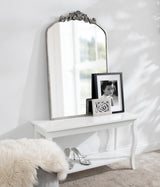Tall arch-shaped wall mirror with ornate carved floral decoration at the top, placed above a white console table holding two framed black-and-white portraits, fluffy beige rug nearby.
