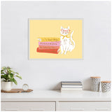 Framed wall art featuring a white cat with pink glasses sitting next to a stack of three books with pastel yellow background and a beaded silver frame.