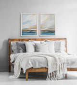 Two framed abstract landscape paintings with soft blue, beige, and gray tones hang above a wooden bed with white and gray pillows and a textured white throw blanket.