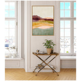 Abstract landscape painting with layers of muted purple, yellow, green, and beige hues in a gold frame hung on a light wall between two tall windows.