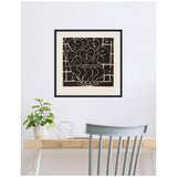 Black and beige framed Henri Matisse print depicting a stylized bowl of begonia leaves with geometric border, hanging above a light wood desk with green plant and blue-gray chair.