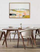 Abstract landscape painting in muted pastels with horizontal bands of pink, yellow, green, and gray, hung above a modern wooden dining table with cream chairs.