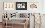 Three framed artworks above a beige sofa with patterned cushions and a chunky knit throw, featuring illustrations of boots, bold lettering 'Howdy Howdy,' and a hat on neutral backgrounds.