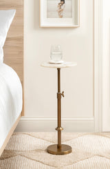 Small round marble-top side table with adjustable bronze metal base, holding a glass of water beside a bed with wood frame and white bedding on textured beige rug.