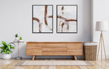 Two abstract canvas artworks with curved brown brushstrokes on white backgrounds, hung side by side above a light wood sideboard in a minimalist living room.