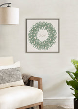 Square canvas wall art featuring a symmetric green wreath with the word 'JOY' centered inside, framed in a simple dark frame hanging above a cream armchair.