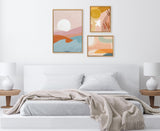 Three framed abstract canvas artworks above a white bed with white pillows and beige blanket, featuring warm pastel hues of orange, pink, blue, and yellow in organic shapes.