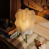 Glowing white wavy-patterned lamp shade on a small tripod stand with green legs, placed on a textured multicolored rug near stacked books and a seashell.