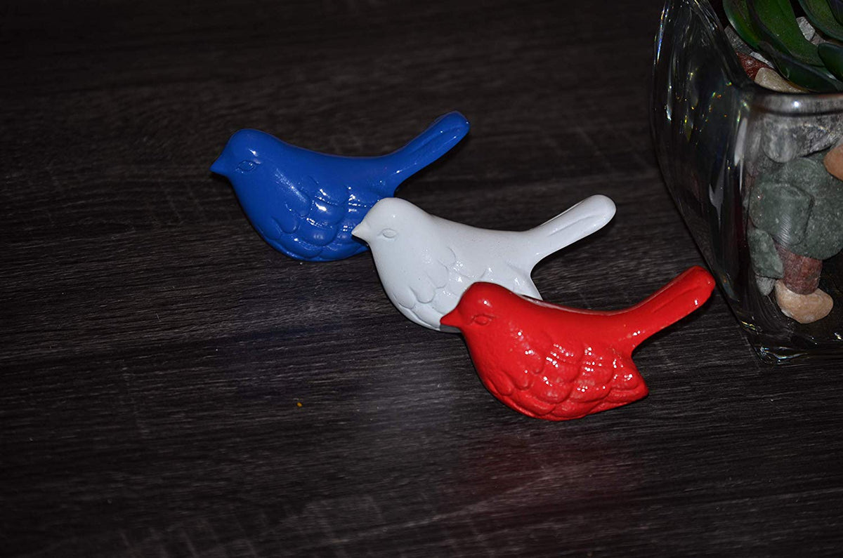 Three small bird figurines in blue, white, and red arranged diagonally on a dark wooden surface near a glass container with green and brown elements.