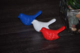 Three small bird figurines in blue, white, and red arranged on a dark wooden surface next to a glass container with stones and a plant.