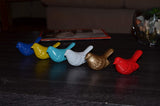 Row of six small bird figurines in blue, yellow, turquoise, white, bronze, and red, lined up on a dark wooden surface.
