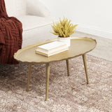 Oval coffee table with scalloped edge and slender tapered legs in matte gold finish, placed on patterned beige rug beside cream armchair with brown knit throw.