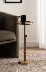 Polished brass adjustable metal side table with a circular top holding a black mug, positioned next to a dark green upholstered armchair on a textured beige rug.