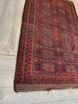 antique 1880s Baluch rug in deep red with geometric patterns and multiple contrasting borders, displayed diagonally on light wood flooring