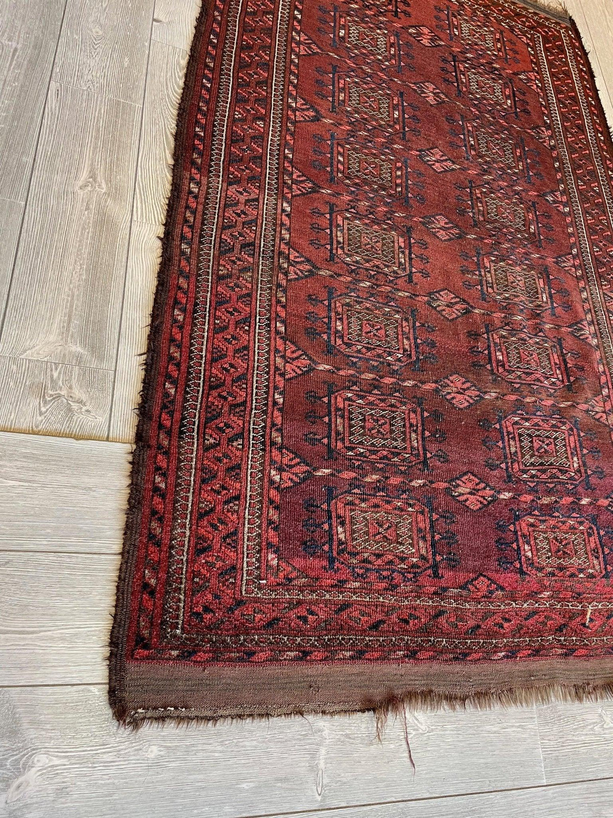 antique 1880s Baluch rug in deep red with geometric patterns and multiple contrasting borders, displayed diagonally on light wood flooring