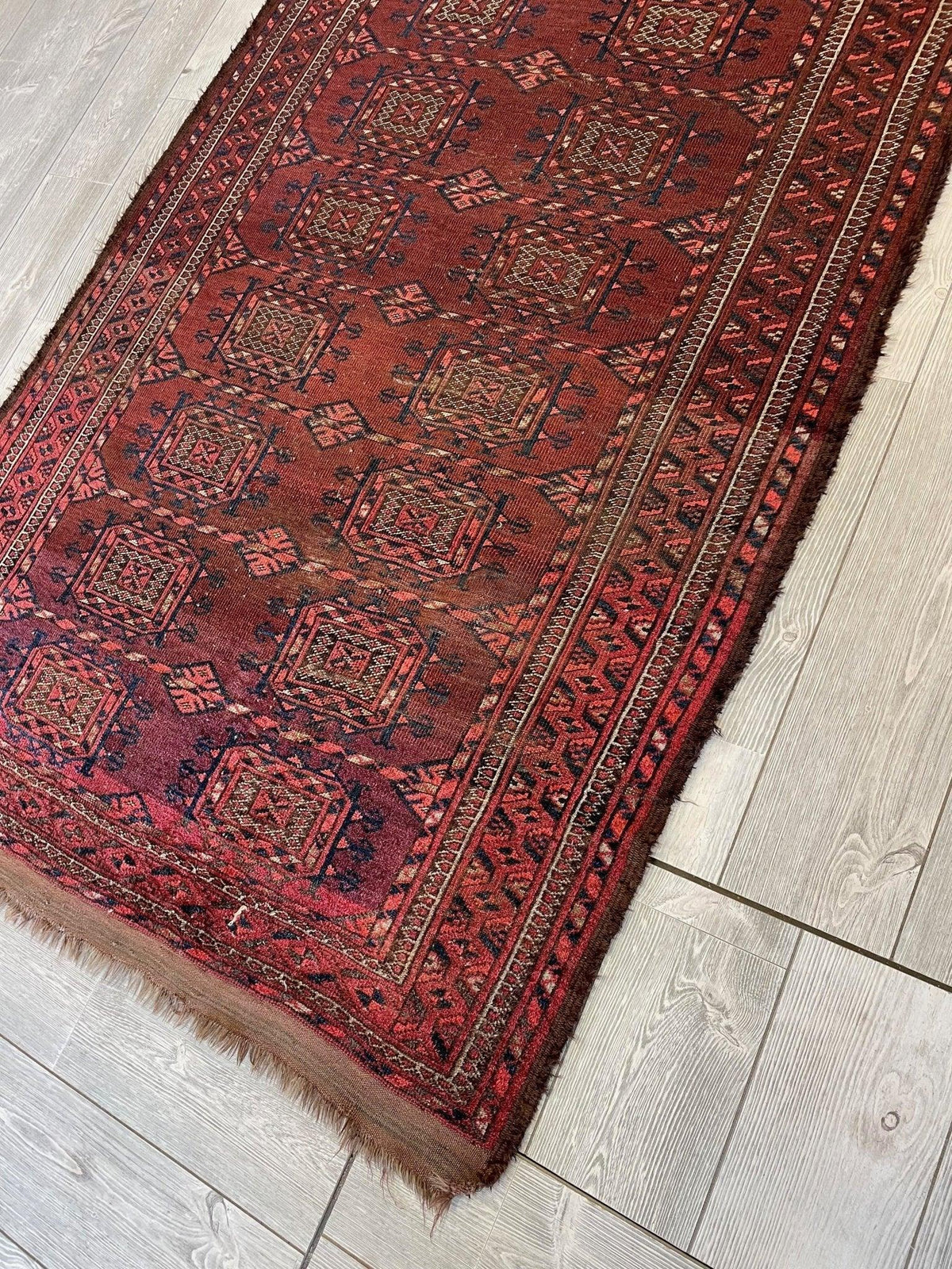 Antique 1880s Baluch rug with deep red and brown geometric patterns framed by multiple patterned borders, displayed on light wood flooring.