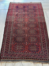 Rectangular antique Baluch rug featuring deep red and dark blue geometric motifs with a detailed border and fringed edges, laid on a wooden floor.