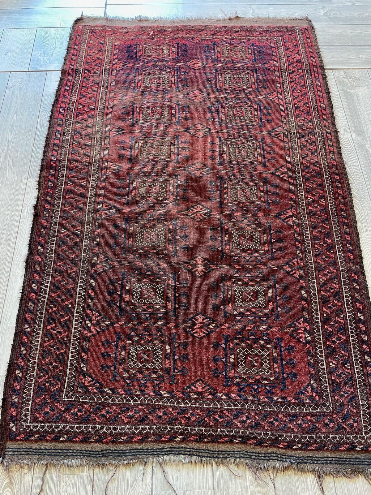 Rectangular antique Baluch rug featuring deep red and dark blue geometric motifs with a detailed border and fringed edges, laid on a wooden floor.