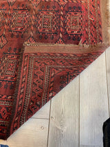 Corner of an antique 1880s Baluch rug with deep red and brown geometric patterns, showing intricate border designs and some frayed edges, lying on light wood floor.