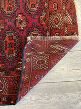 Antique 1910s Turkoman rug in deep reds with geometric medallions and tribal motifs, showing a folded corner revealing intricate reverse patterns on light wood flooring.