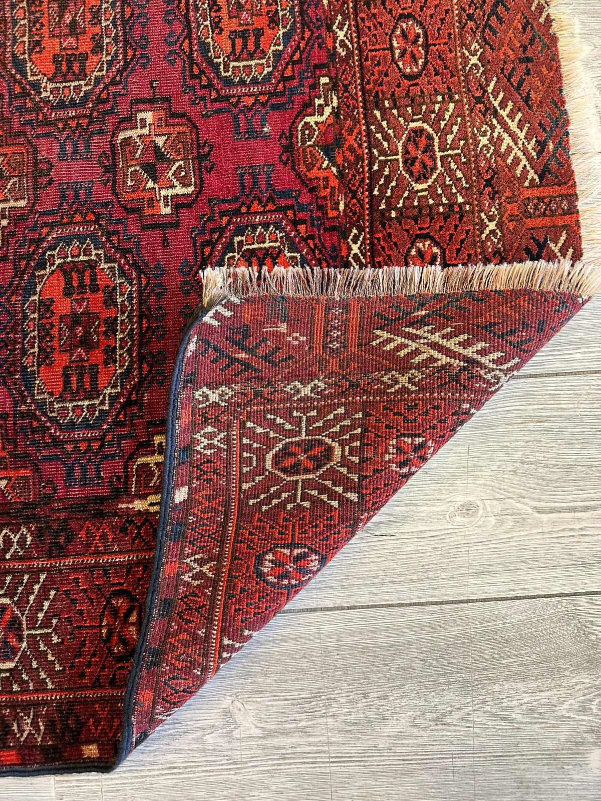 Antique 1910s Turkoman rug in deep reds with geometric medallions and tribal motifs, showing a folded corner revealing intricate reverse patterns on light wood flooring.
