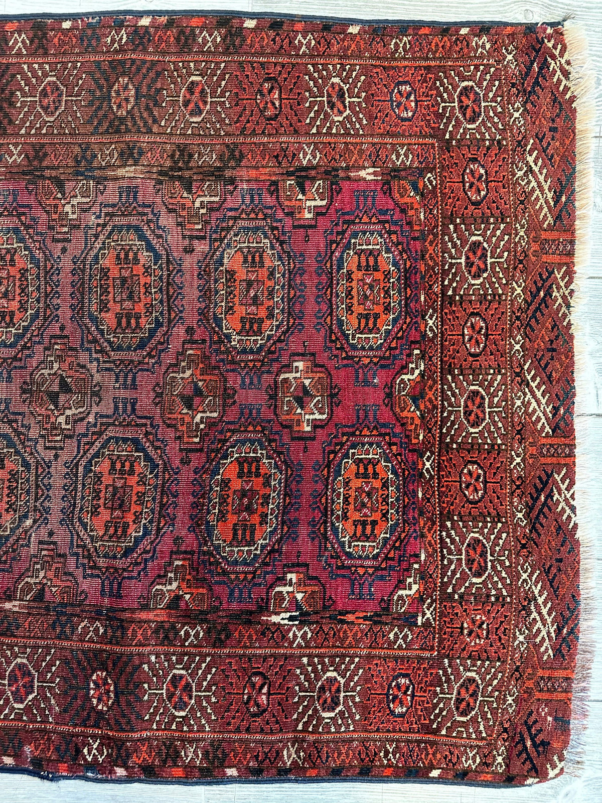 Rectangular antique Turkoman rug featuring repeating geometric medallions in deep red and navy on purple background, bordered by red bands with star motifs and white fringe.