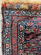 Close-up of an antique Kurdish rug corner showing intricate floral and geometric patterns in red, blue, cream, and black with a worn border and knotted fringe.