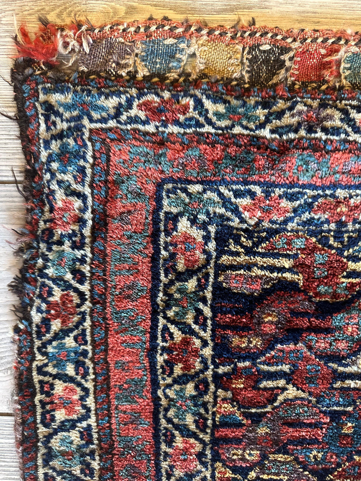 Close-up of an antique Kurdish rug corner showing intricate floral and geometric patterns in red, blue, cream, and black with a worn border and knotted fringe.