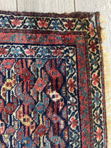 Corner of a small antique Kurdish square rug displaying intricate geometric and floral patterns in red, blue, cream, and black with fringed edge on wooden floor.