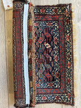 Small folded Kurdish square rug with rich red, blue, and cream geometric and floral patterns, showing frayed edges and a measuring tape for scale.