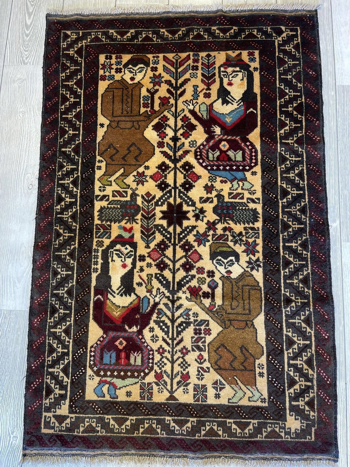 Rectangular Baluchi rug with two pairs of stylized human figures facing a tree, framed by intricate dark geometric borders on a light brown field.
